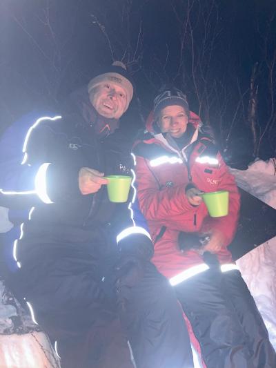 a man and woman in snow gear holding cups around a campfire 